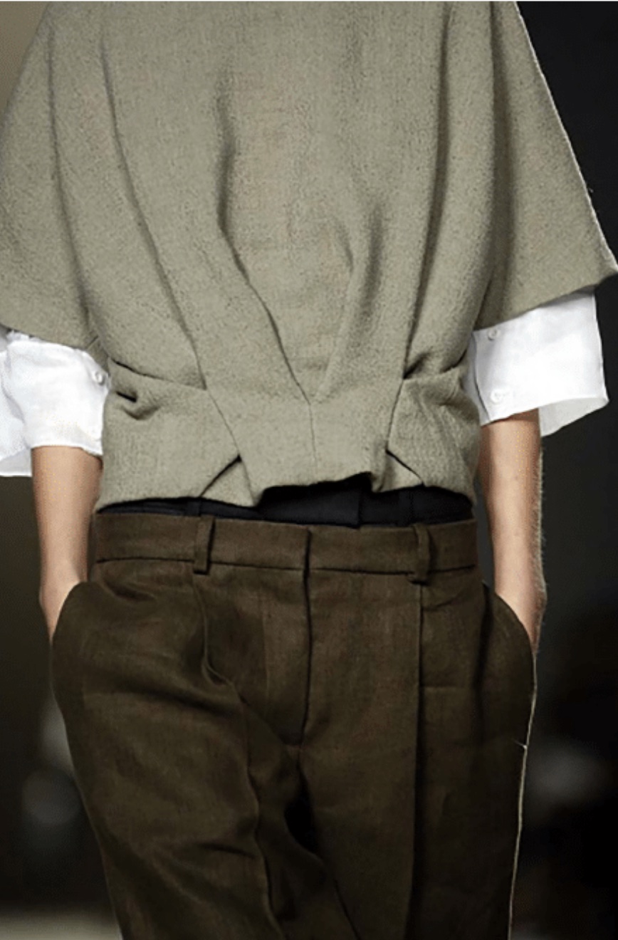 A mid body shot of a chloe model with their hands in their pockets, wearing brown suit trousers and three quarter, bell sleeved shirt and jumper.